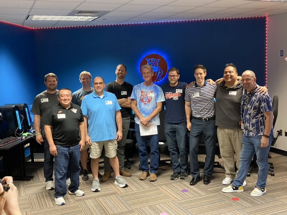 Group of ten Blackland Prairie Wildcat dads standing in a blue-lit room with a Wildcat logo on the back wall. They are gathered for Fifth Grade TEKS Day to teach about density.