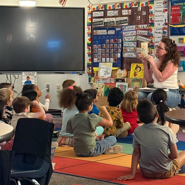 A teacher reads a book to a group of young, diverse elementary students sitting on a brightly colored rug in a classroom.