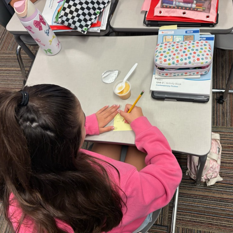 An overhead view of a student with long brown hair writing on a yellow sticky note with a pencil.
