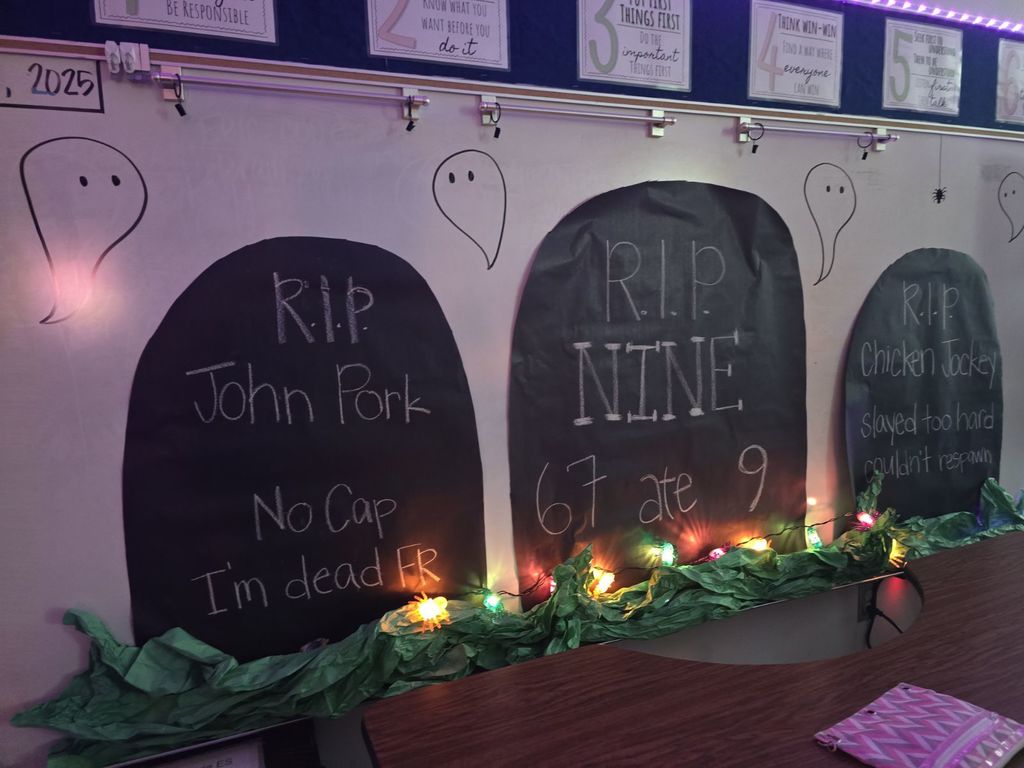 A classroom whiteboard decorated for Halloween with three large, black paper tombstones featuring humorous, student-written epitaphs like "R.I.P. NINE / 6 7 ate 9."
