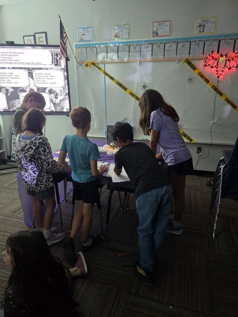 A group of elementary students gathers around a table with a purple cloth and electric candles, working on papers in a dimly lit classroom decorated with caution tape.