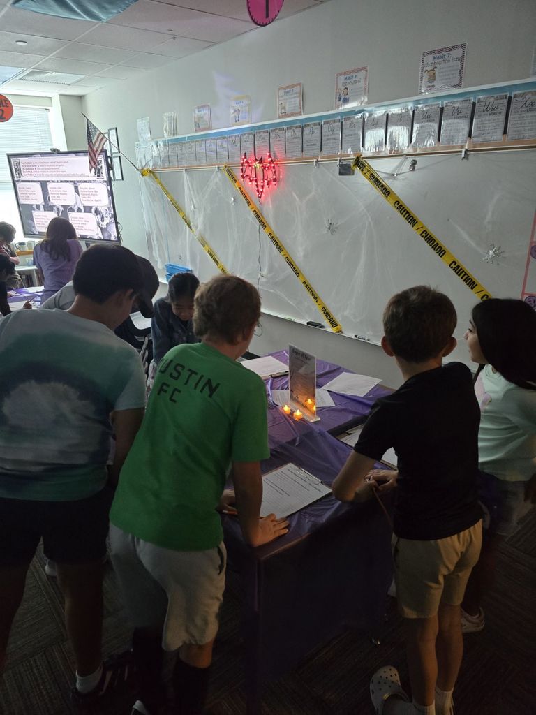 A group of elementary students gathers around a table with a purple cloth and electric candles, working on papers in a dimly lit classroom decorated with caution tape.