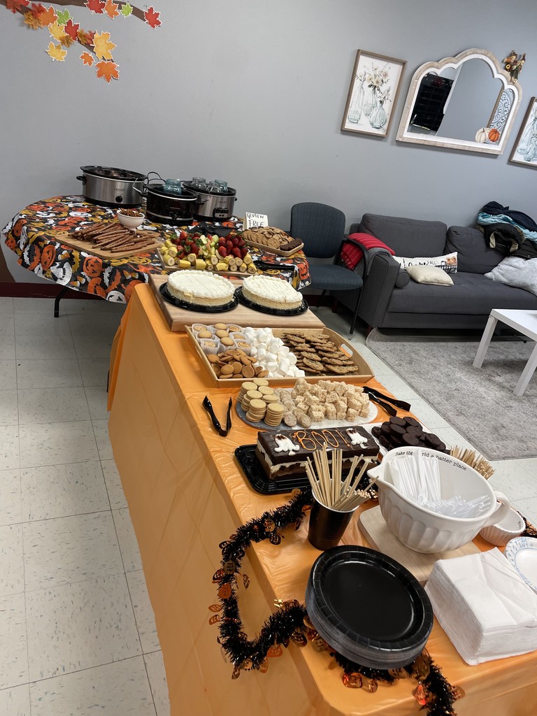 A close-up shot of a food table decorated with a Halloween-themed tablecloth. The table holds three slow cookers, a tray of chocolate-covered pretzel rods, a large fruit tray with bananas and strawberries, and a plate of cookies. In the foreground, two large cheesecakes sit on a separate, orange-covered table.