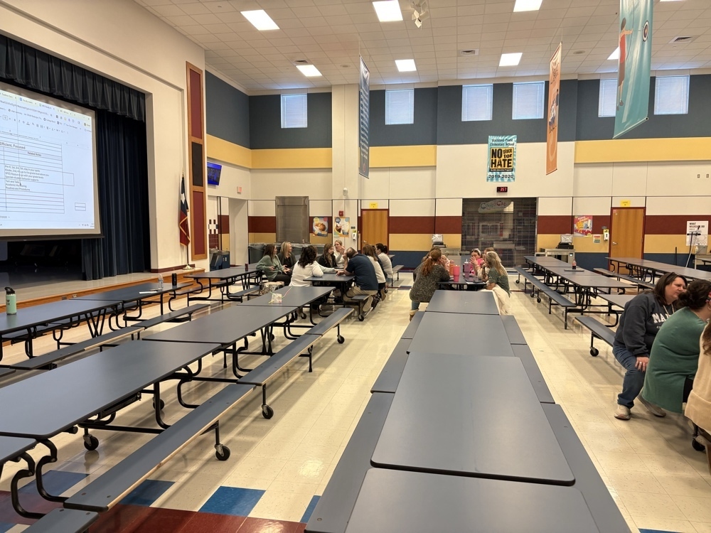 A wide view of the school cafeteria showing staff members seated at tables during a professional development session. A large projection screen at the front of the room displays an organized spreadsheet titled Efficient, Focused, which outlines grade level schedules and procedures.