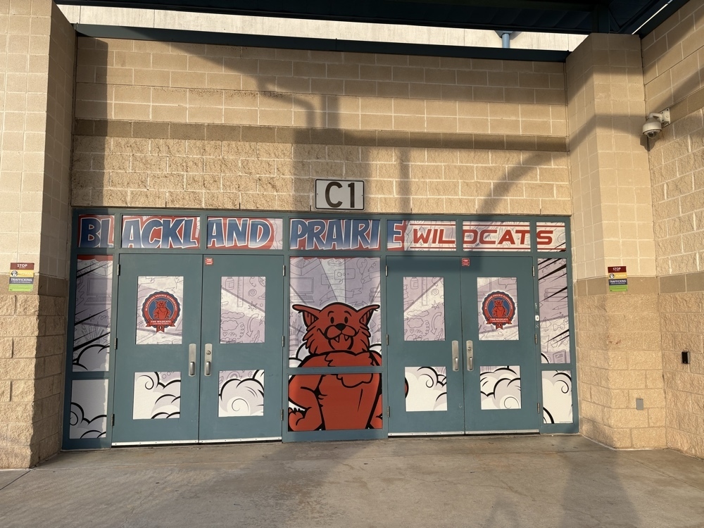 A wide view of the school entrance labeled C1 showing four blue doors and surrounding windows covered in school branded wraps featuring the Blackland Prairie Wildcats mascot and logo.