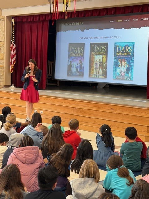 Alyson Gerber standing  in front of screen, in front of students sitting on the floor
