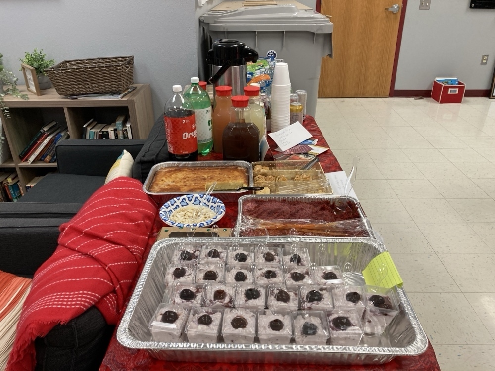 A table covered in a red tablecloth holds several trays of desserts and beverages. The spread includes small individual cups of a creamy dessert topped with a cherry, several trays of traditional sweets, and various drinks including tea, soda and juice.