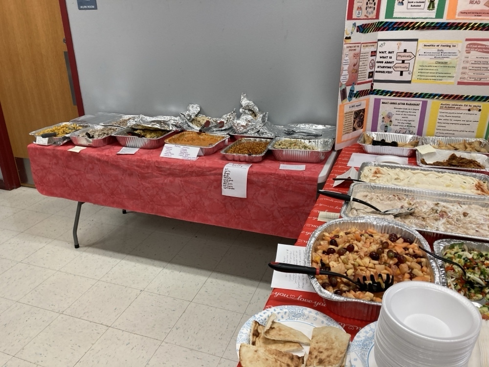 A long buffet line features various savory dishes in aluminum trays, including rice, curry, and flatbreads. A large tri-fold poster board stands at the end of the table, displaying educational information about the traditions and significance of Ramadan.