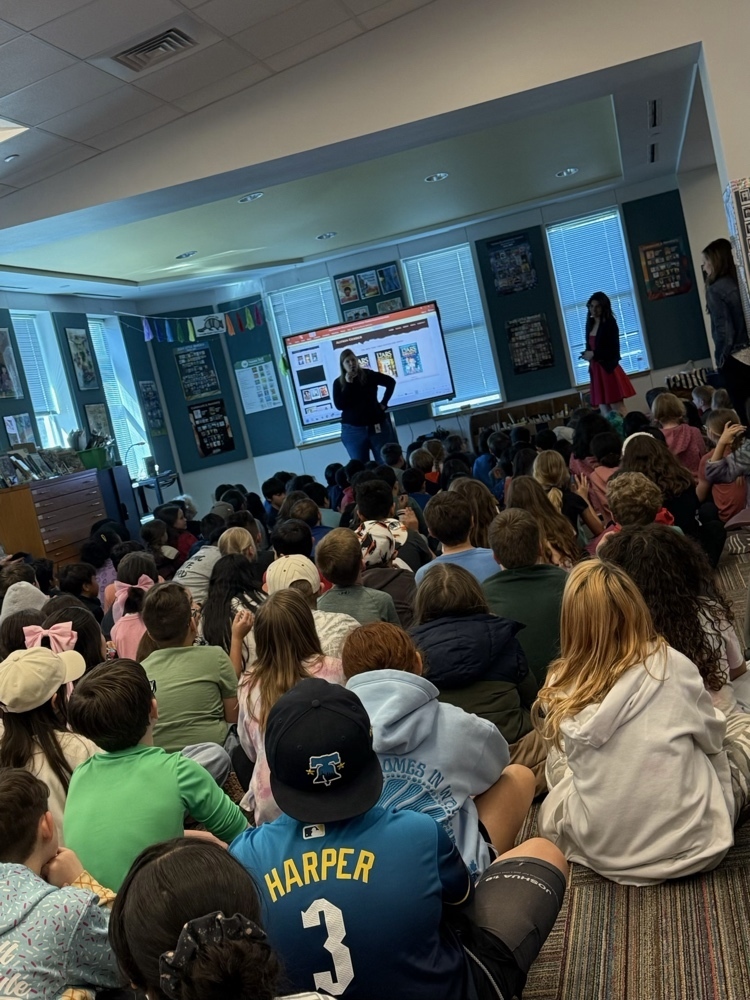A wide shot of a large group of elementary students sitting on the floor of the school library. They are facing a large screen that displays the website of author Alyson Gerber. Two staff members stand near the screen addressing the attentive audience.