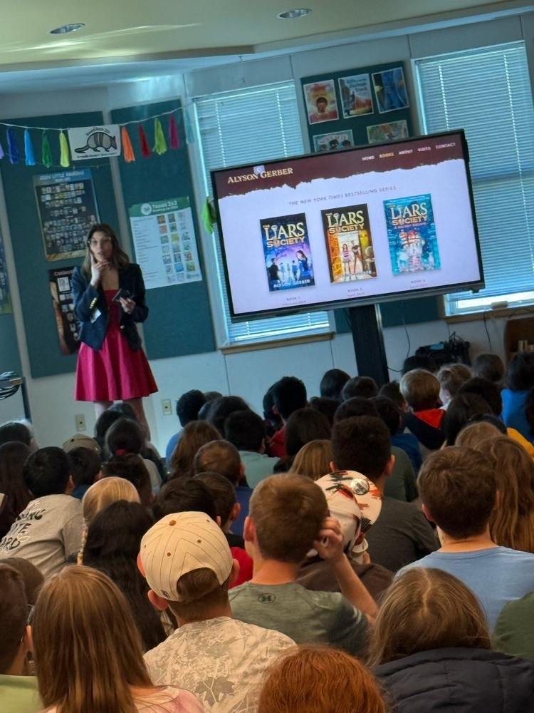 A close up shot of author Alyson Gerber speaking to students in the Blackland Prairie library. She is standing next to a large monitor displaying the covers of her book series, The Liars Society. The room is decorated with educational posters and colorful banners.