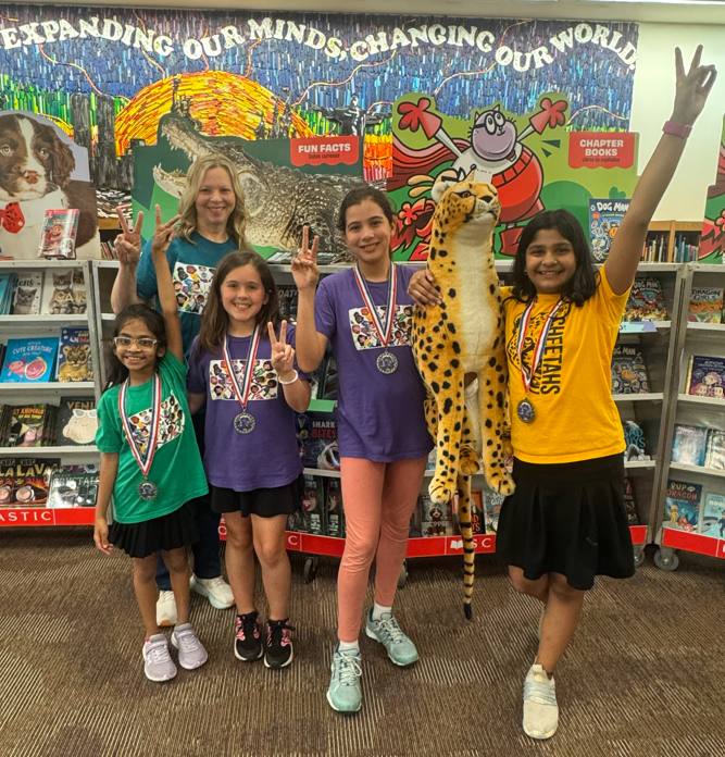 Four students and the librarian standing in front of shelves of books wearing medals and holding a stuffed cheetah- Holding up two fingers