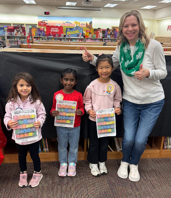 Three Pre-K Students holding a coloring sheet standing in front of black paper, standing next to our Librarian