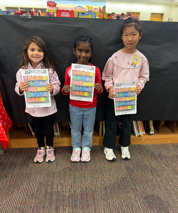 Three Pre-K Students holding a coloring sheet standing in front of black paper