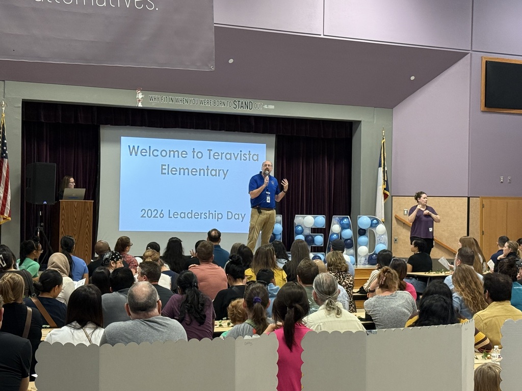 A wide shot of a presentation in a school auditorium or cafeteria. On a stage, a male speaker in a blue shirt and khaki pants gestures while speaking into a microphone. Behind him, a large screen displays the text: "Welcome to Teravista Elementary, 2026 Leadership Day." Large light-blue and white balloons spelling "LEAD" stand to his right, and a woman provides sign language interpretation on the far right. The foreground is filled with the back of a seated audience.