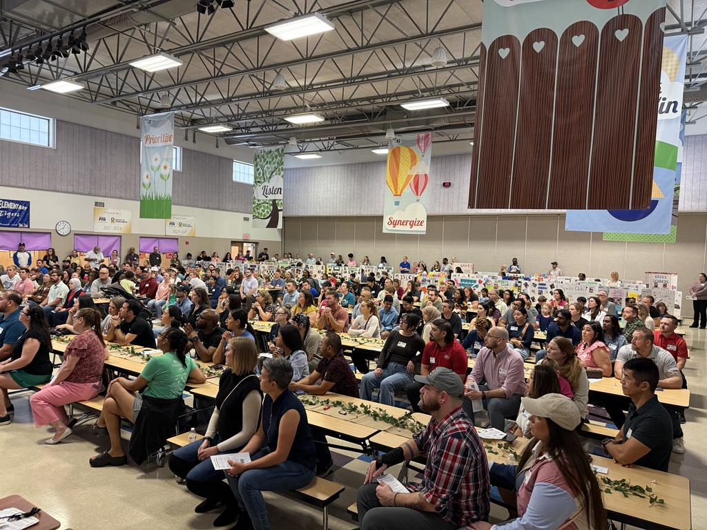 A high-angle view of a large, diverse crowd seated at long cafeteria tables in a school multipurpose room. The audience is facing forward, presumably toward a stage. Colorful banners hang from the ceiling with leadership-themed words like "Prioritize," "Listen," and "Synergize." In the background, various student project boards are lined up against the wall, and the room is brightly lit by overhead industrial lighting.