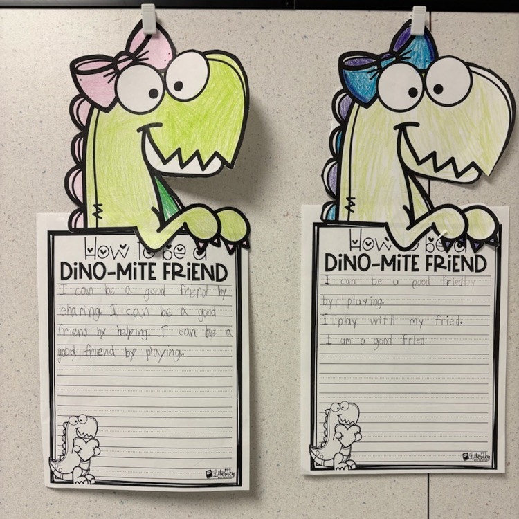 Two student writing projects about being a good friend are displayed, each topped with a colorful dinosaur craft wearing a bow.