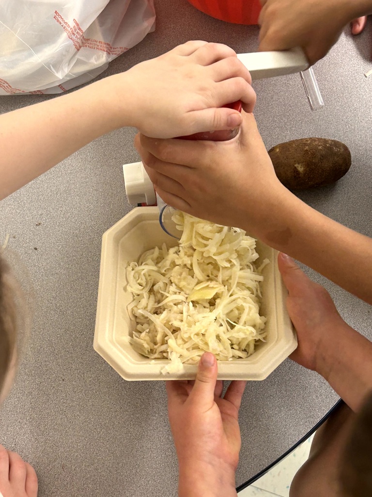 Several students work together to use a manual shredder to turn a fresh potato into a bowl of shredded hash browns.
