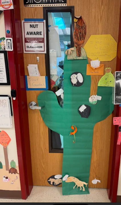 Photograph of a cactus and animals made out of paper on a door. A folder on the door that says: Tour Guides, with papers in it. Sign that says Warning! Wach out Saguaro Cactus. This is how it others know.  An orange envelope that says: Fun Facts, with notecards inside