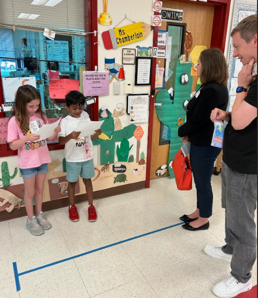 Two students reading from papers standing in front of wall with cacti and two parents standing by listening