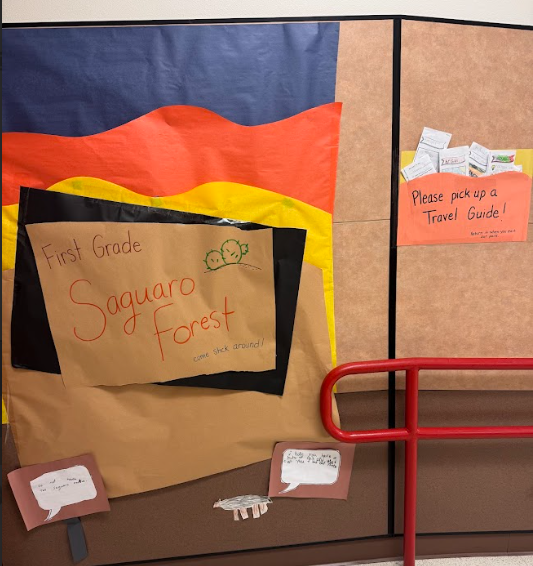Photograph of colored paper on the wall that says "First Grade Saguaro Forest" and has a folder with papers that says "Please pick up a Travel Guide!"