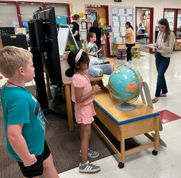 Two students reading from papers standing in front of  board and table with a globe, and adults and another student standing