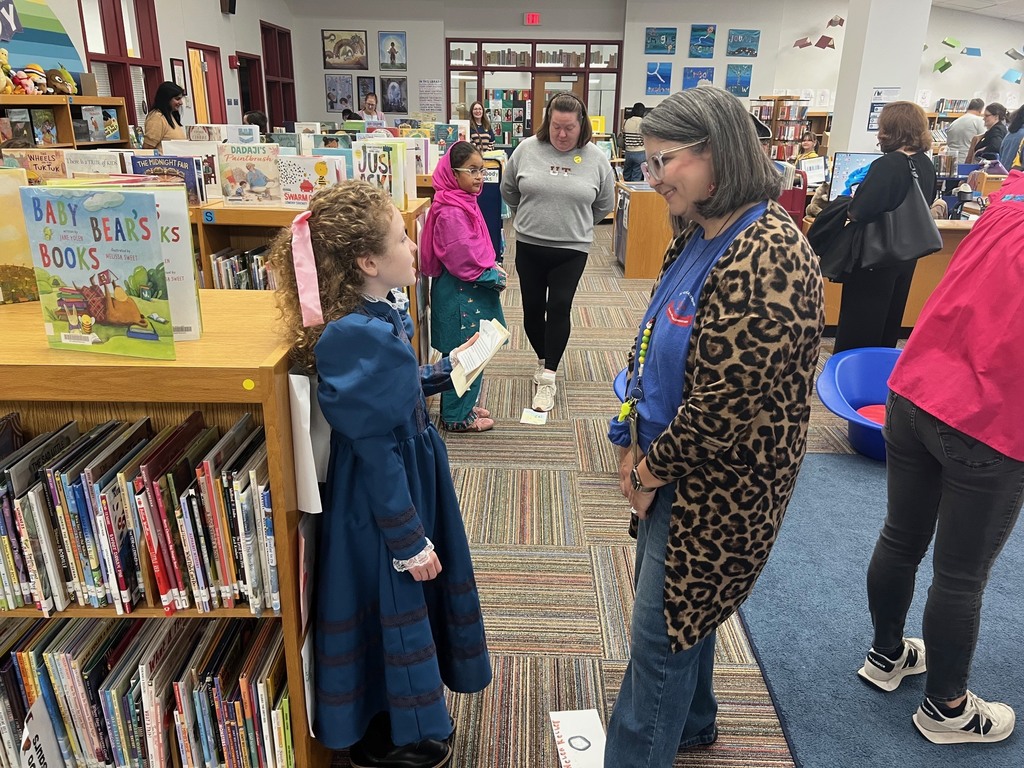 A student dressed in a long blue historical gown and pink hair bow speaks to a visitor among the library bookshelves.