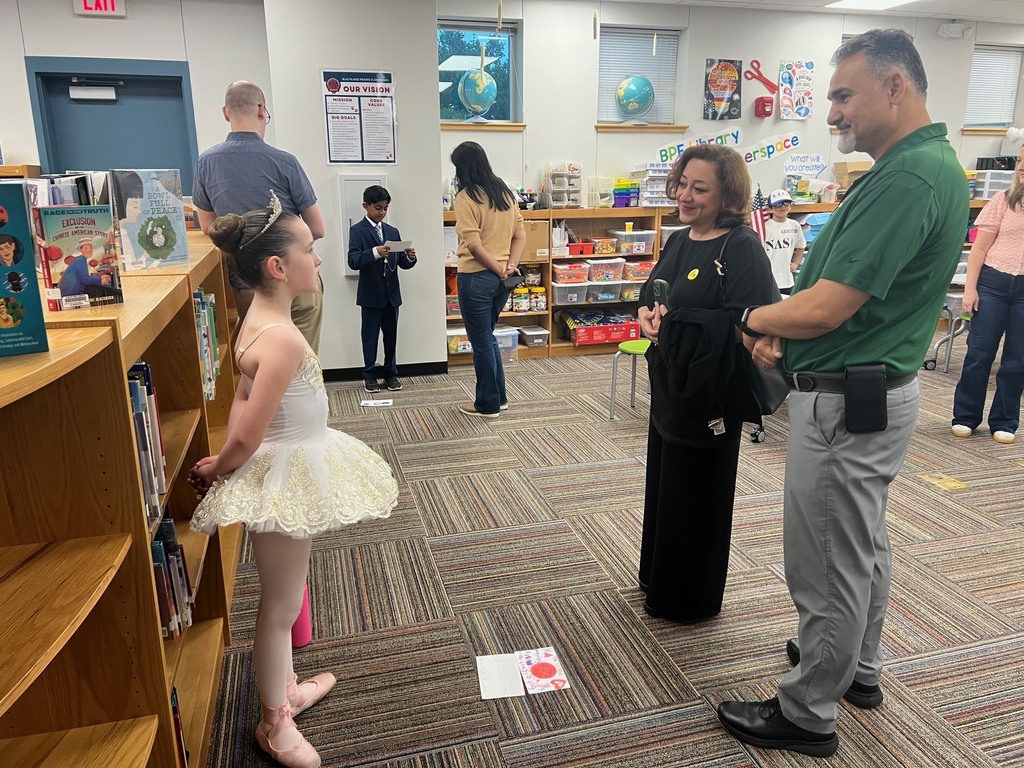 A student dressed in a white ballet tutu and tiara stands near a bookshelf while presenting her Living Wax Museum project to two adults.