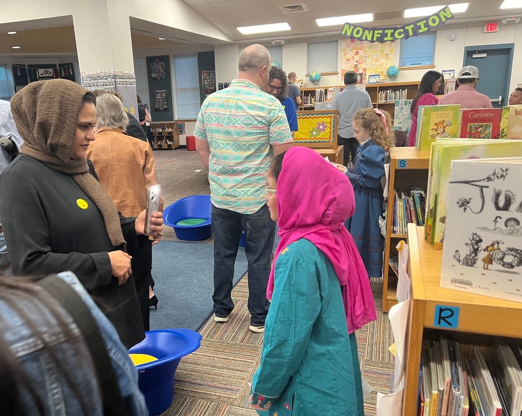 A third grade student in a bright pink headscarf and teal outfit presents her historical research to a parent in the school library.