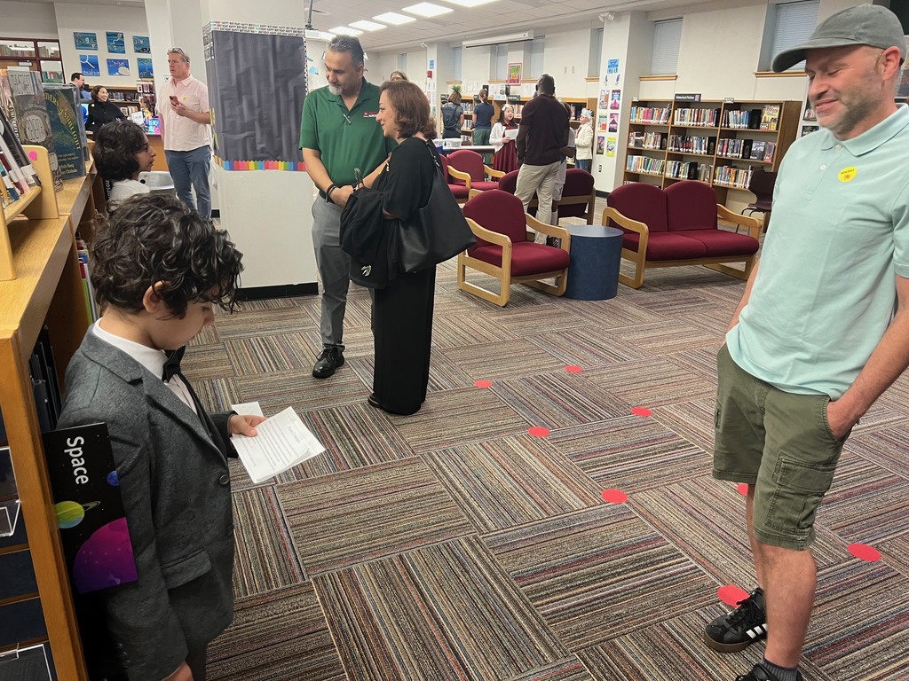 A student in a grey suit jacket holds a script and prepares to present his project to a visitor standing nearby in the library.