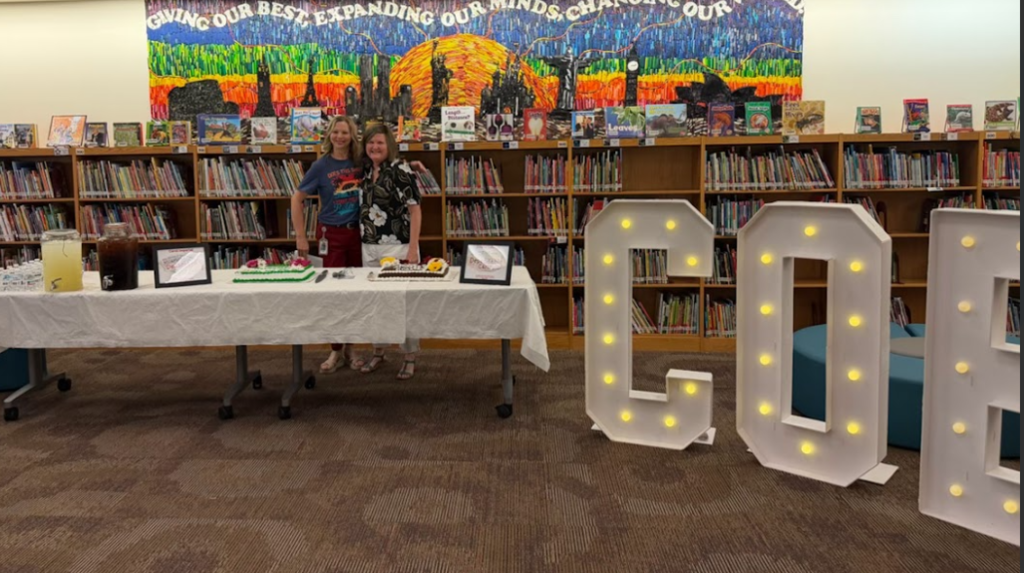 Two women standing in front of bookshelves in the library, standing behind a white table with two cakes and picture frames, and drinks. Standing next to the giant lit up letters COE