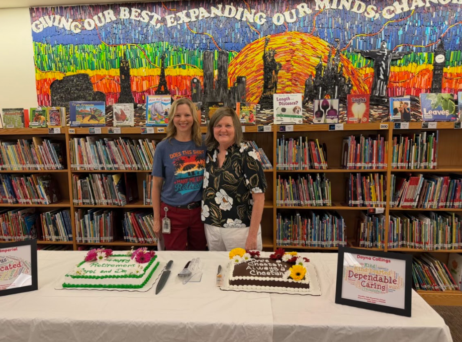 Two women standing in front of bookshelves in the library, standing behind a white table with two cakes and picture frames. 