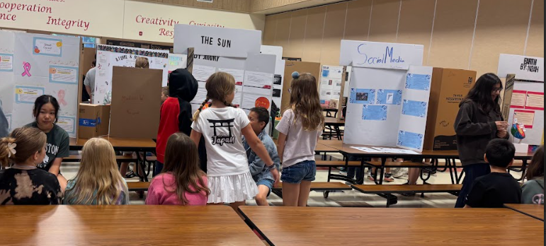 students sitting at a table watching other students present in front of their trifold boards set on a table. 