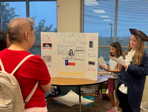 Photograph of parents looking at two students presenting next to their trifold board sitting on a table. The trifold board is titled "Exploration & Mission"