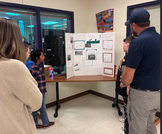Photograph of parents looking at three students presenting next to their trifold board sitting on a table. The trifold board is titled "Cattle Boom"