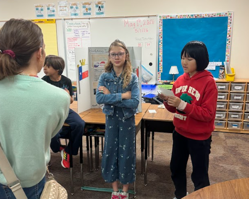 Parent standing in front of three students presenting in front of trifold board sitting on a desk