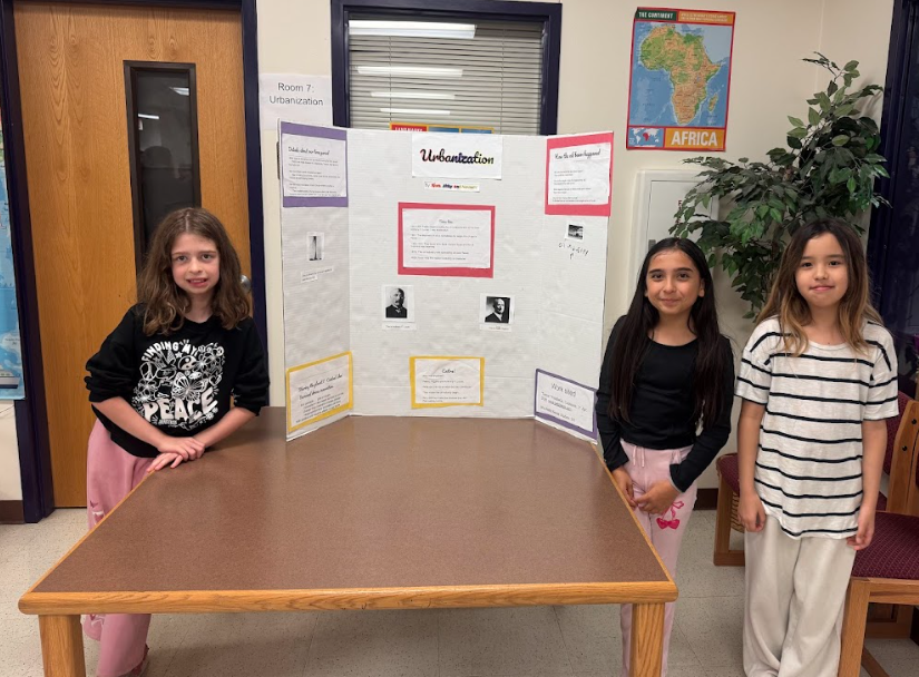 Three students standing next to a trifold board titled "Urbanization" sitting on a table