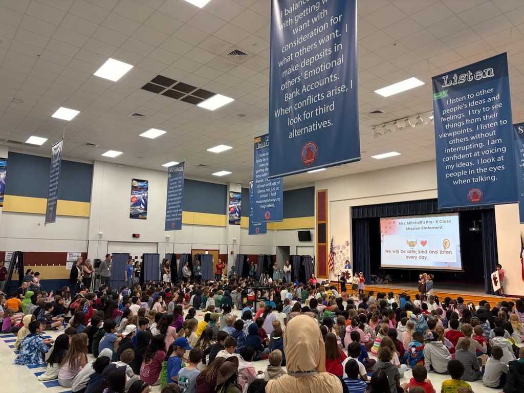 Students sit on the floor of the cafeteria for the Habit Rally. A projector screen shows a Pre-K mission statement while large banners with leadership habits hang from the ceiling.