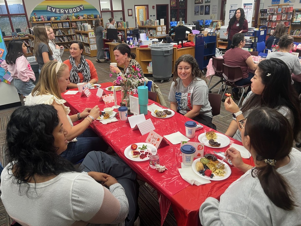 Volunteers sit at a long table with a red tablecloth during the Volunteer Breakfast in the library. They are enjoying food and conversation surrounded by flowers and thank you cards.