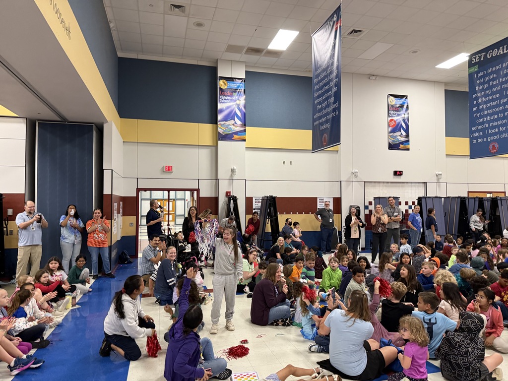 A student stands in the center of the gym during the Habit Rally holding colorful streamers. Staff and students clap and cheer from the sidelines and the floor.