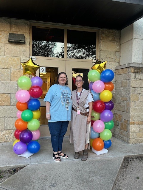 Two teachers standing in front of doors of the school with ballon arches on either side with mutli colored ballons and gold stars on top