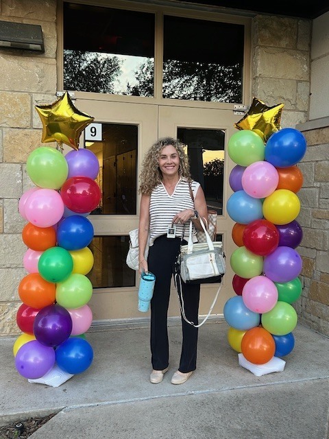 A teacher standing in front of doors of the school with ballon arches on either side with mutli colored ballons and gold stars on top, holding a bag and water cup