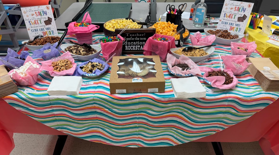 Table covered in different snacks to make trail mix with two signs that say "A little something salty and a little something  sweet because having staff like you is a really big treat"