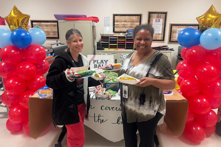 Two teachers standing holding hotdogs, pickles, and snacks in front of table. There are red  two balloon towers on either side with a gold star on top