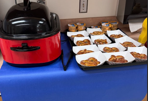 Red hotdog warmer sitting on blue paper with paper containers holding soft baked pretzels with small containers of cheese