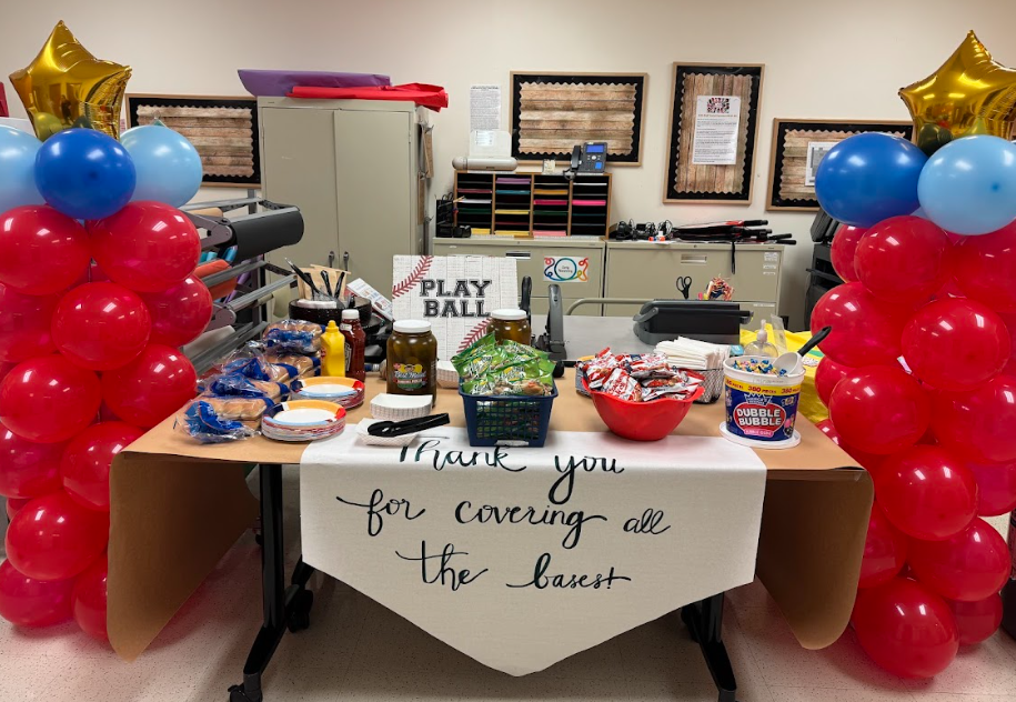 Photograph of table covered in buns, pickles, cracker jacks, peanuts, bubble gum, and plates with a sign that says "Play Ball" and another Sign that says "Thank you for covering all the bases". There are two balloon towers with red balloons, and a gold star on top