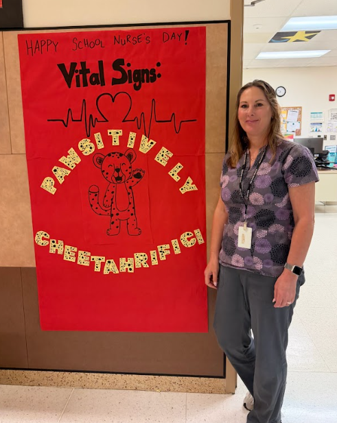 Our School Nurse standing next to a red sign that says "Happy School Nurses Day! Vital Signs: Pawsitively Cheetahrific!" with a cheetah drawn on the sign. 