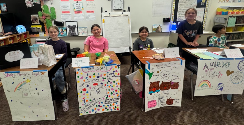 5 students standing behind desks with signs, selling items for the 3rd Grade Economic Fair