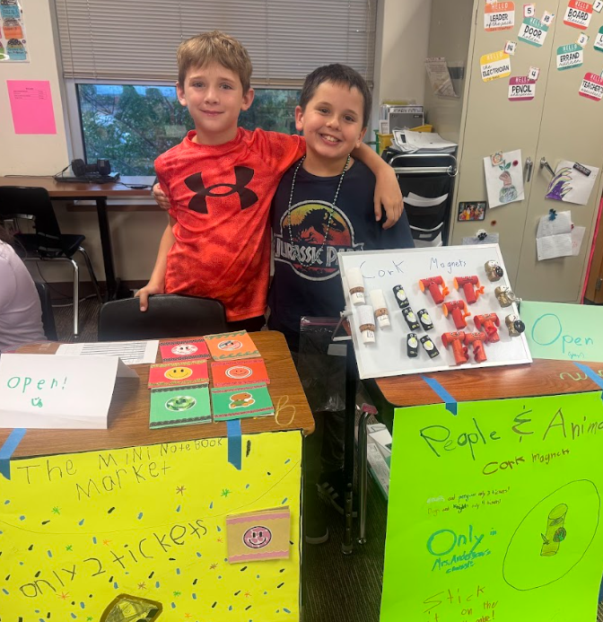 Two students standing behind desks with signs, selling items for the 3rd Grade Economic Fair