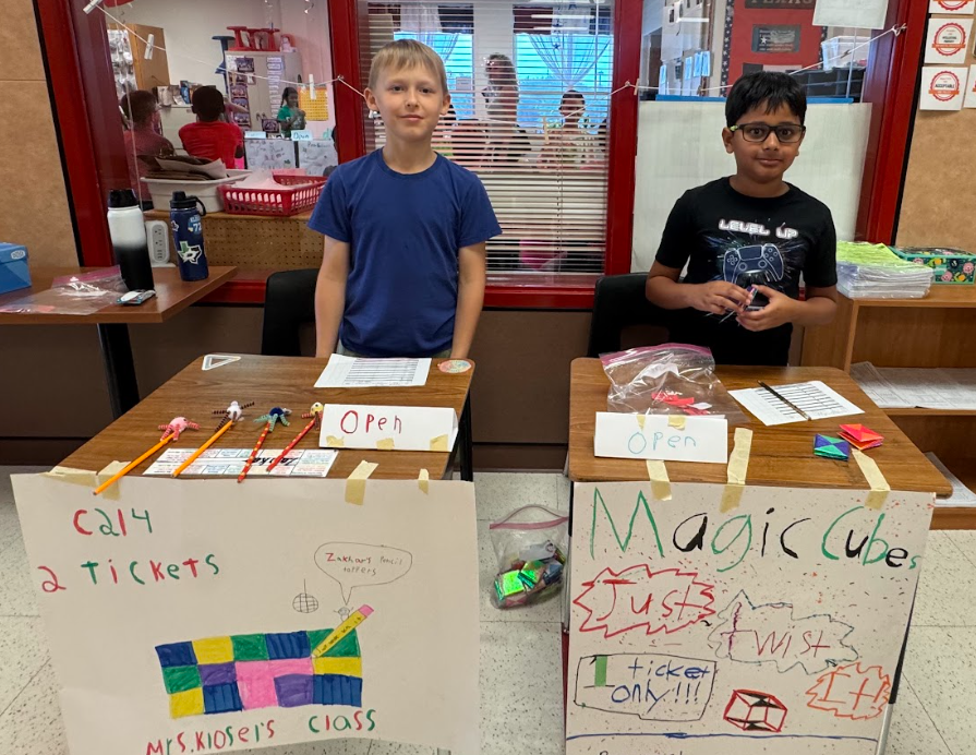 Two students standing behind desks with signs, selling items for the 3rd Grade Economic Fair
