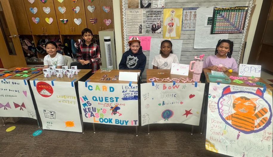 5 students standing behind desks with signs, selling items for the 3rd Grade Economic Fair